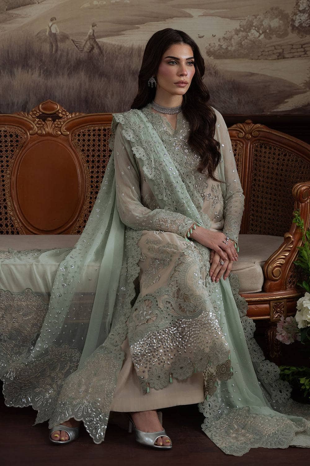 Woman in a light green embroidered outfit sitting on a decorative chair with a scenic background.