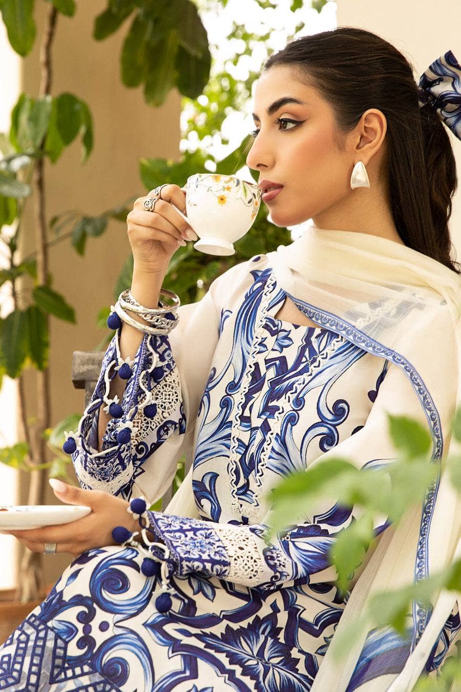 Woman in a blue and white patterned dress drinking from a teacup outdoors.