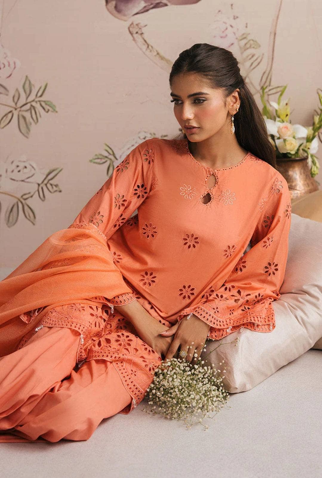 Woman in an orange traditional outfit holding flowers, sitting on a couch with a decorative background.