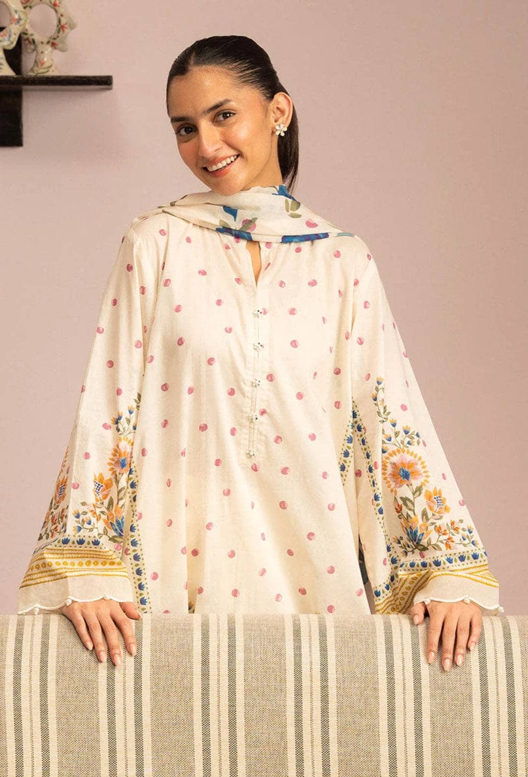 Woman wearing a cream-colored traditional outfit with floral embroidery, sitting on a striped chair against a plain background.