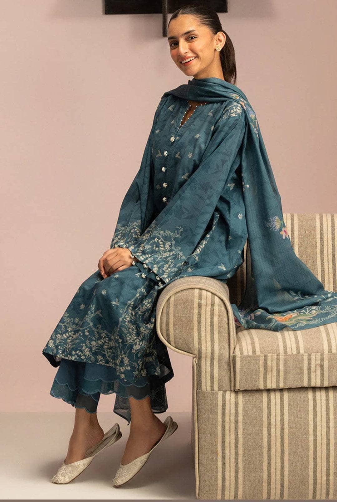 Woman in a blue traditional outfit sitting on a striped chair against a pink wall.