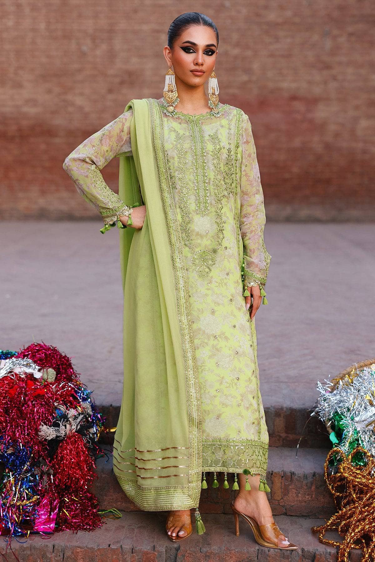 Woman in a green traditional outfit with floral patterns, standing against a brick wall.