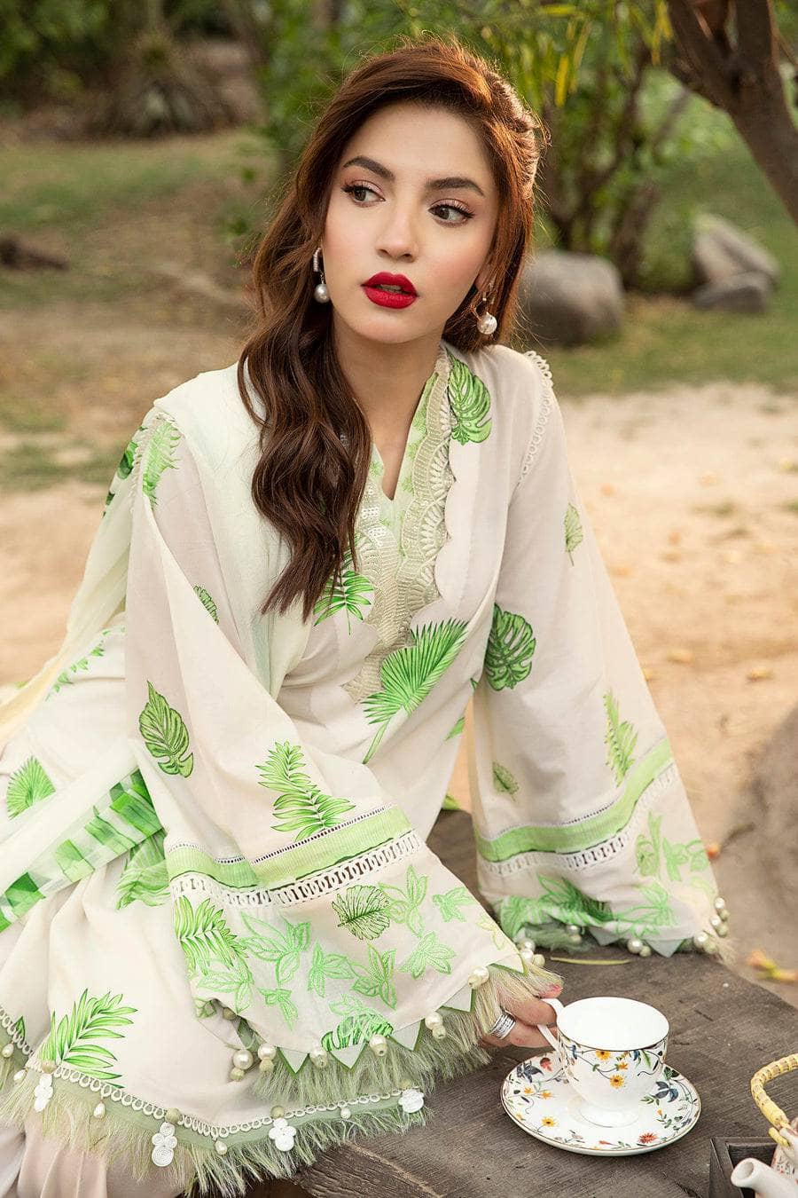 Woman in a white and green patterned outfit sitting outdoors with a cup of tea.