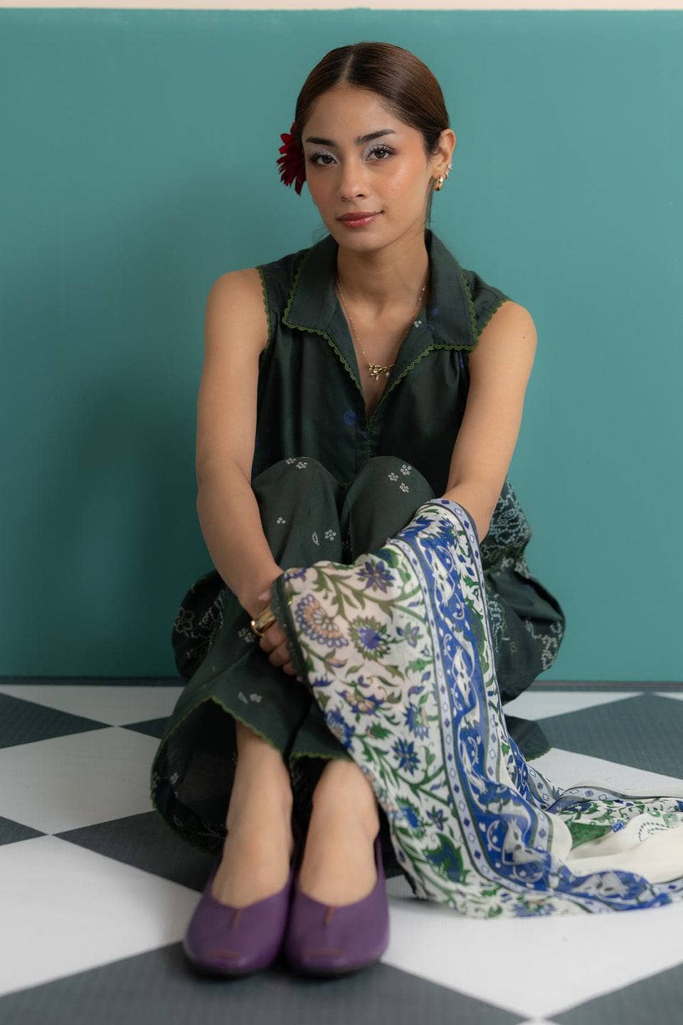 Woman sitting on a checkered floor with a patterned scarf, wearing a green dress and purple shoes.
