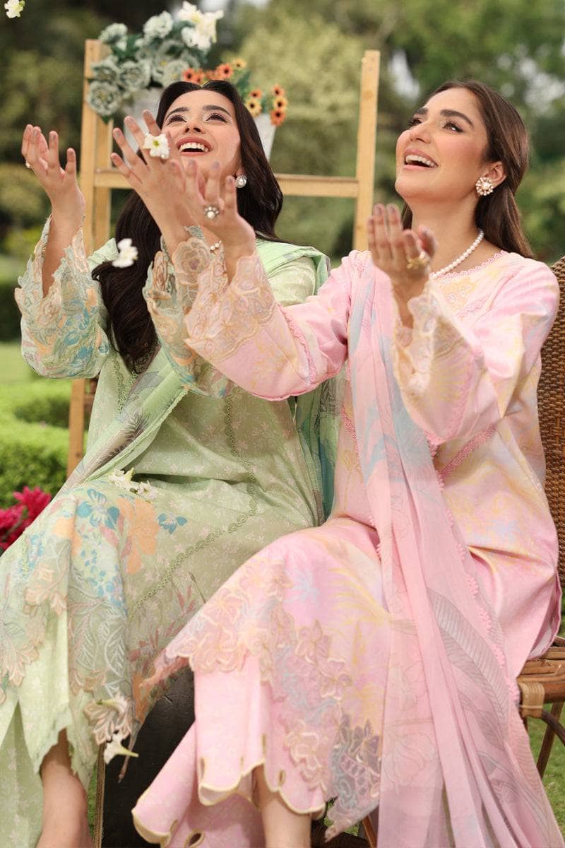 Two women in traditional outfits sitting outdoors with a floral arrangement in the background.