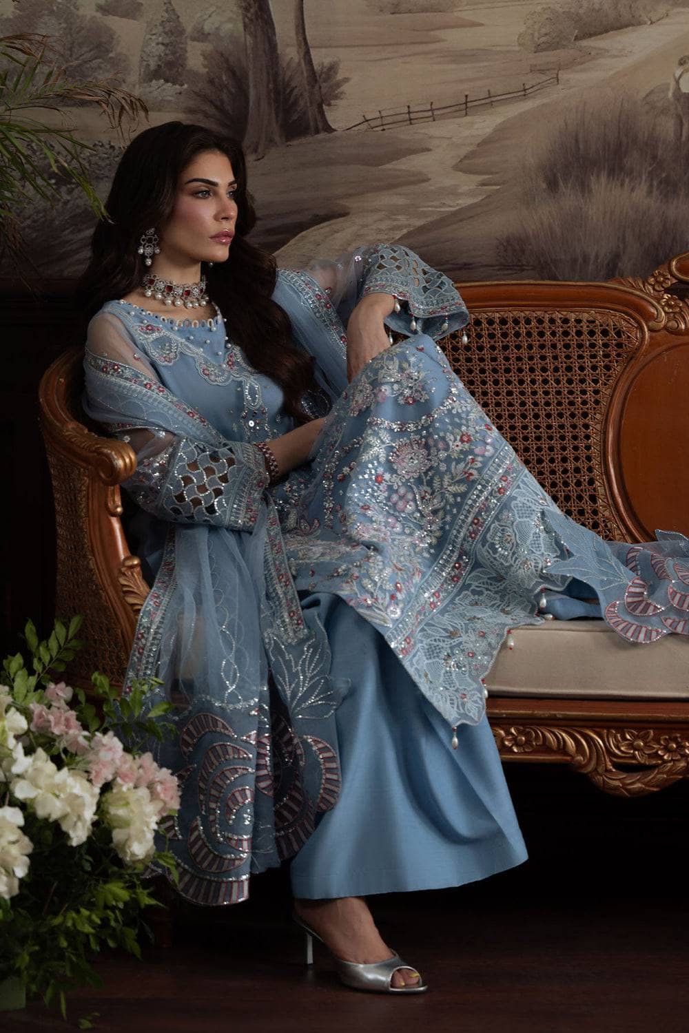 Woman in a blue embroidered outfit sitting on an ornate chair with floral decorations.