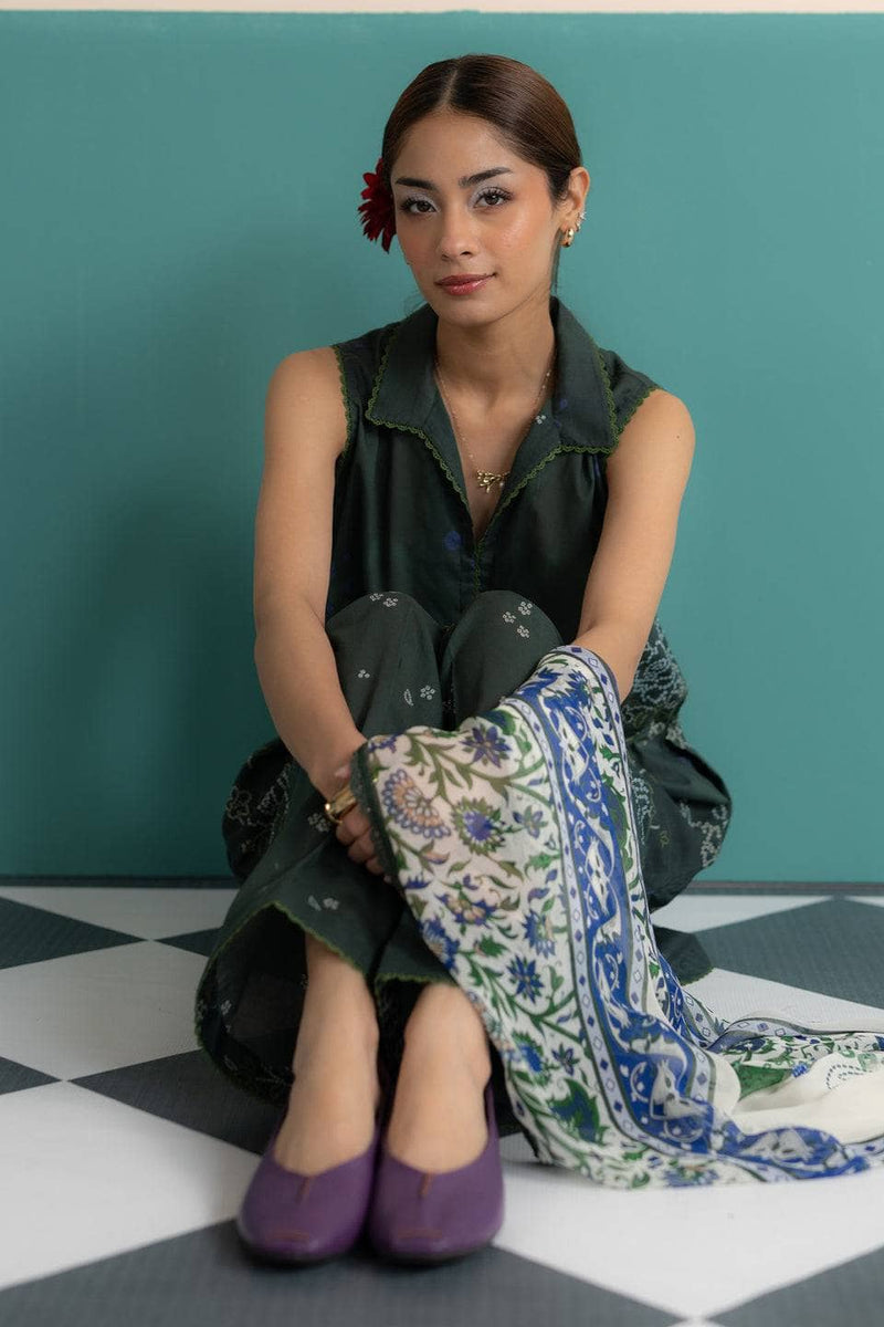 Woman sitting on a checkered floor with a patterned scarf, wearing a green dress and purple shoes.