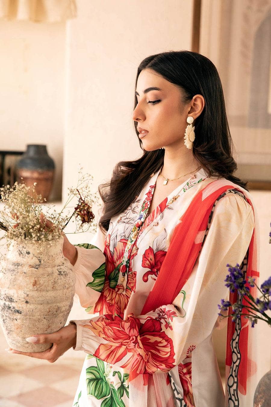 Woman holding a vase with flowers, wearing a floral dress and red scarf.