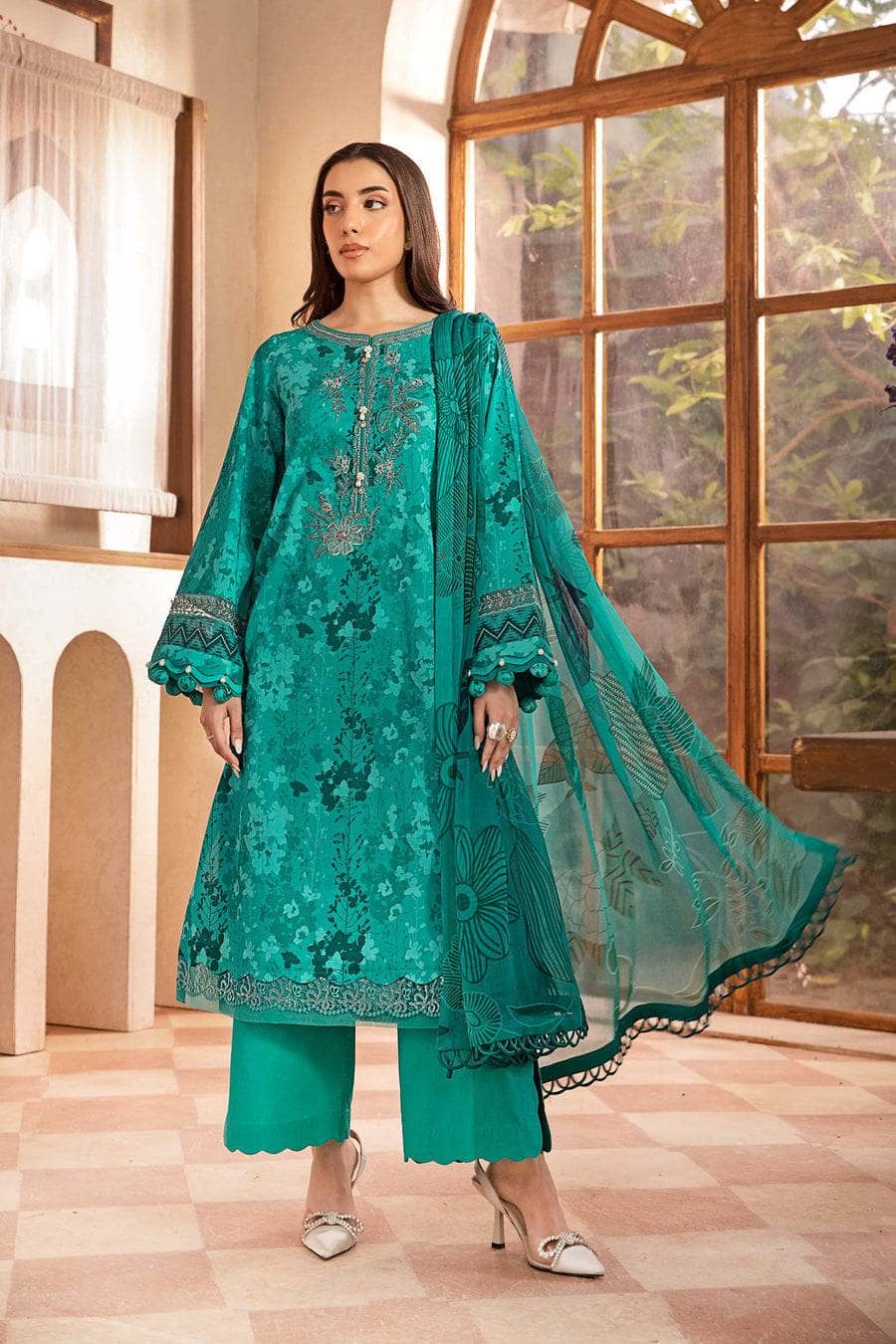 Woman wearing a teal embroidered outfit in a room with large windows.