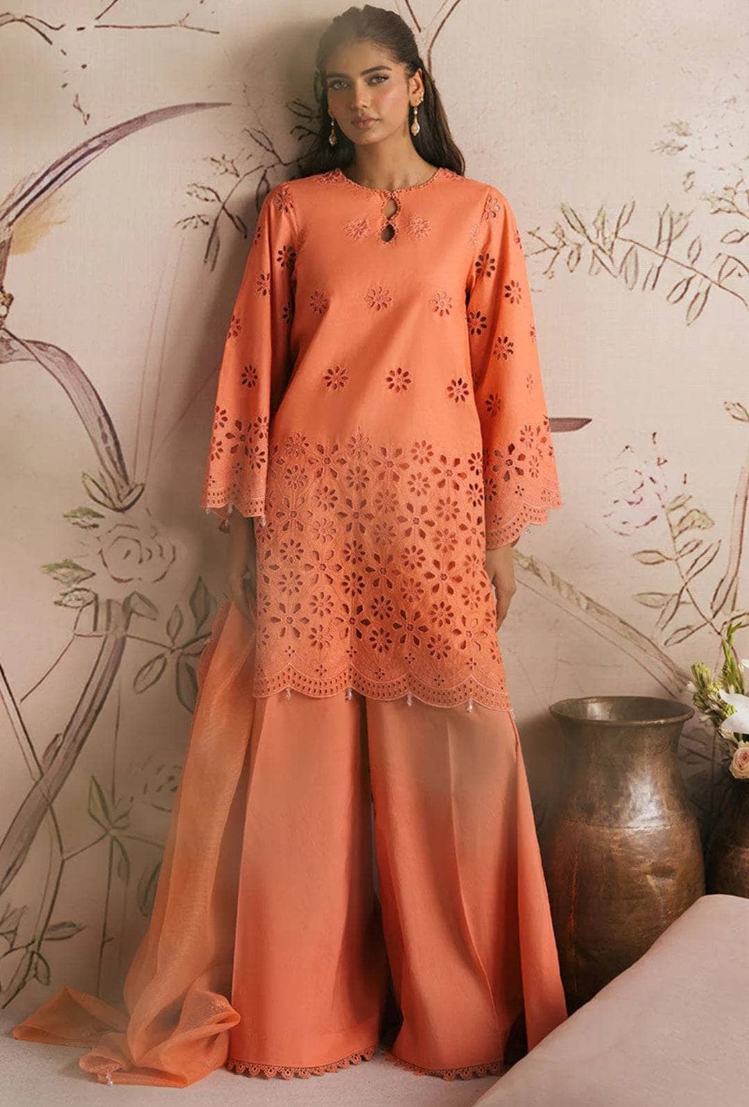 Woman wearing an orange embroidered outfit against a decorative wall.