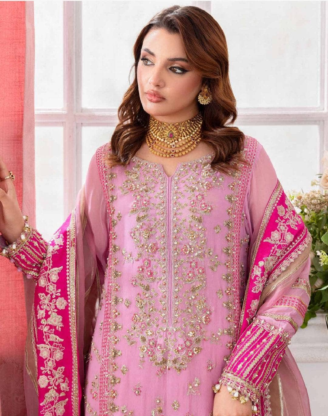 Woman wearing a pink embroidered traditional outfit with gold jewelry indoors.