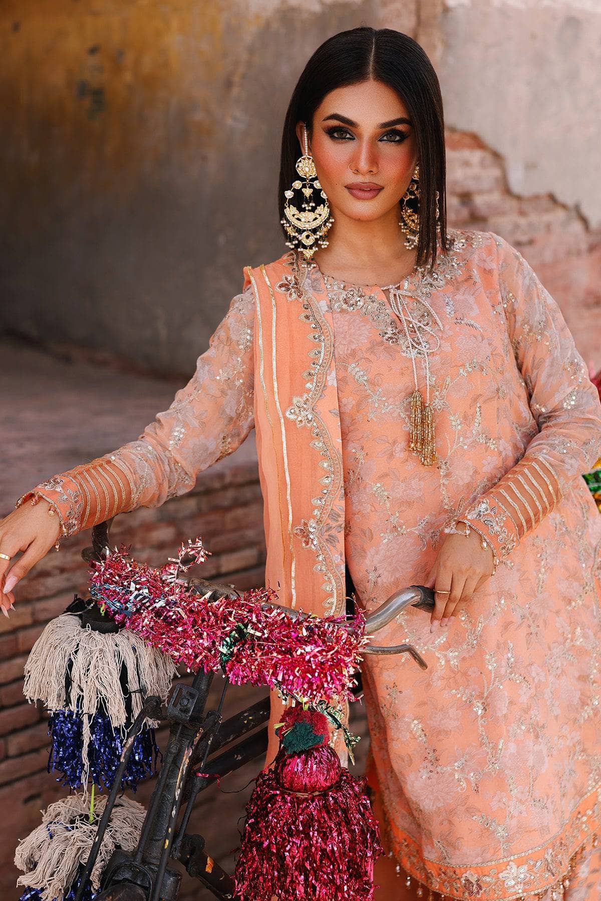 Woman in a peach embroidered outfit holding flowers, standing against a textured wall.