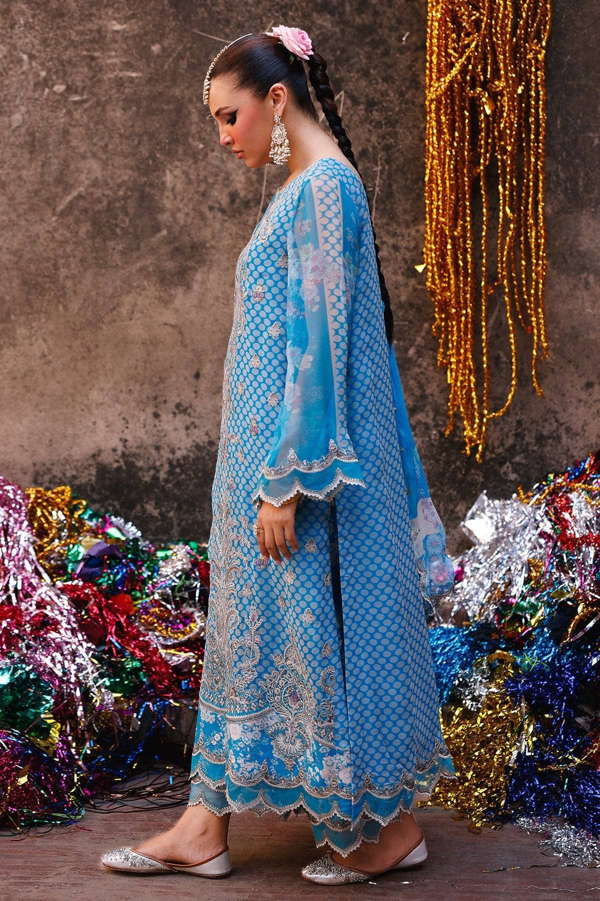 Woman in a blue embroidered traditional outfit standing against a textured wall with decorative ribbons.