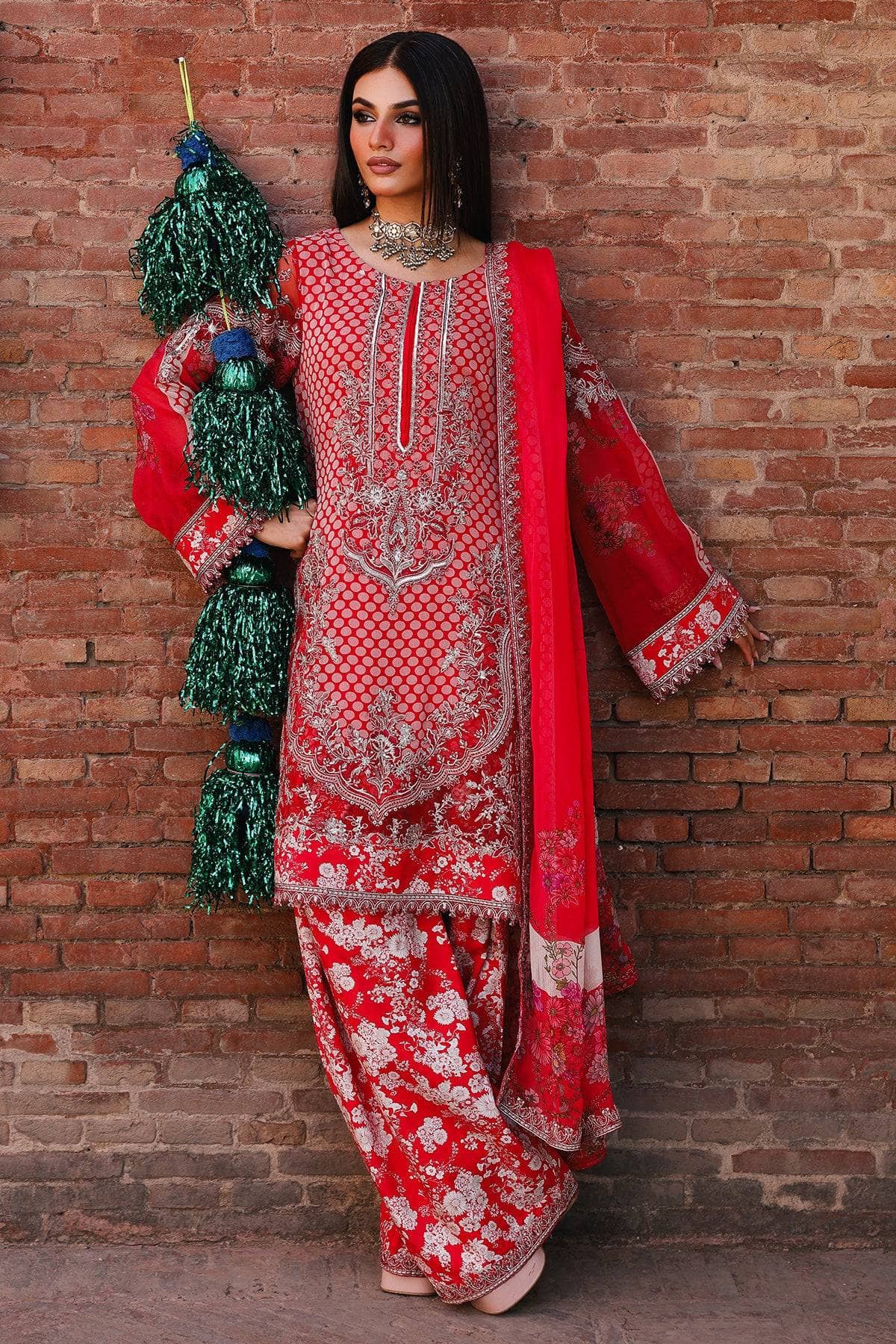 Woman in a red and white embroidered traditional outfit holding green decorative items against a brick wall.