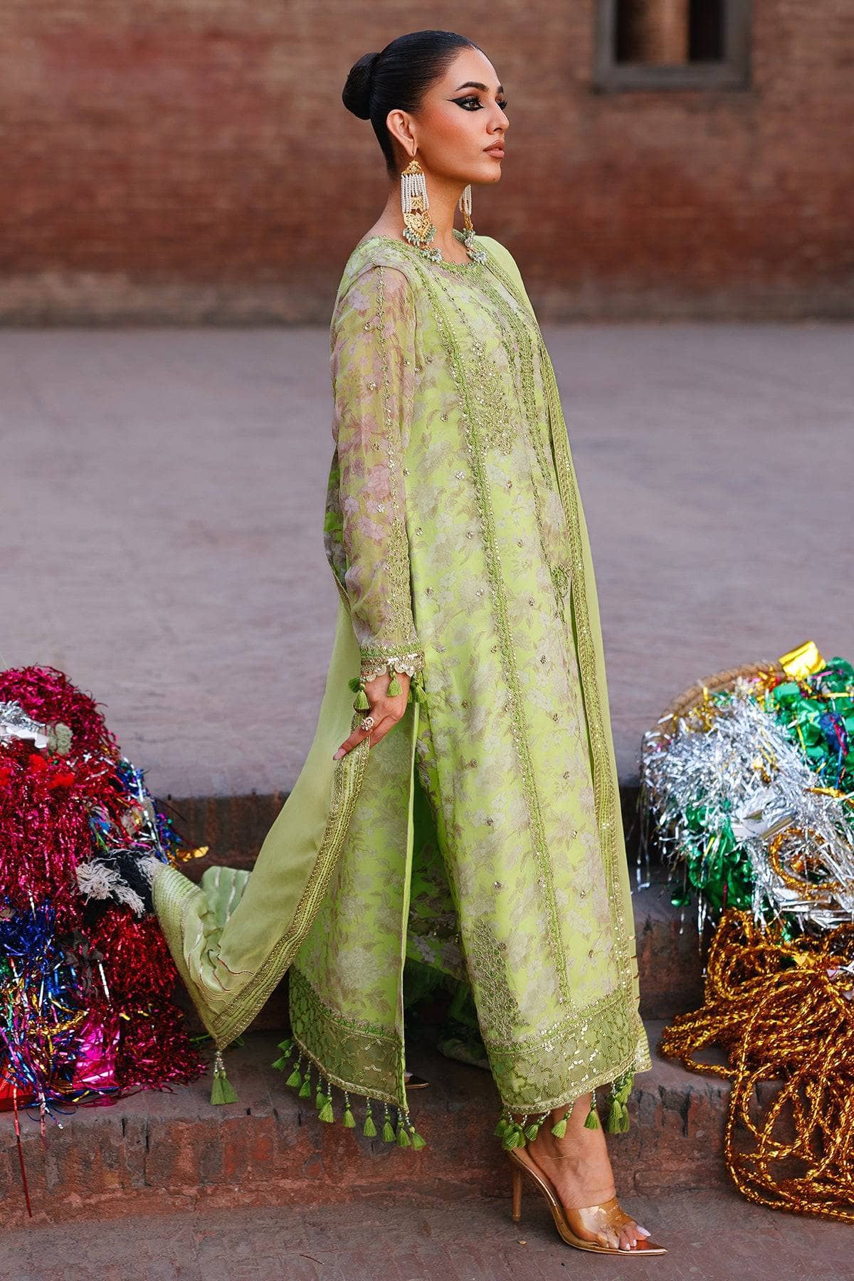 Woman in a green traditional outfit standing in front of decorative items against a brick wall.