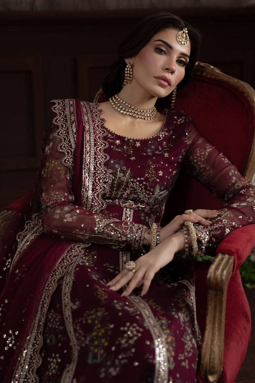 Woman in a maroon embroidered traditional outfit sitting on a red chair.
