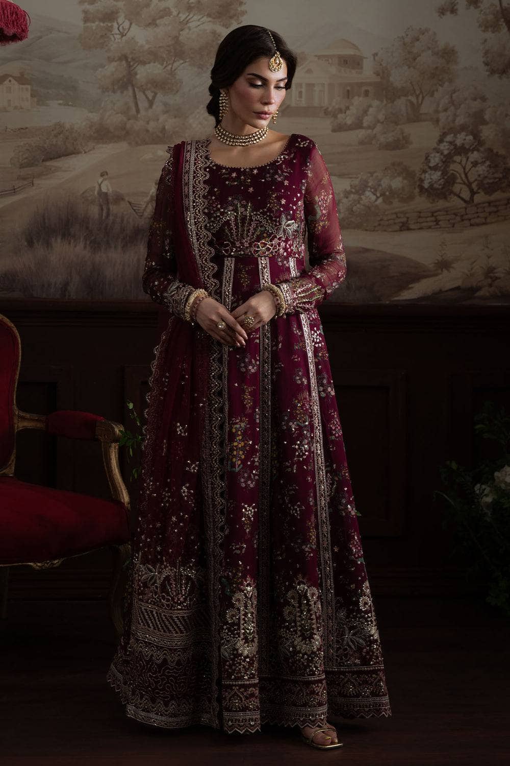 Woman in a maroon embroidered traditional outfit standing against a decorative wall.
