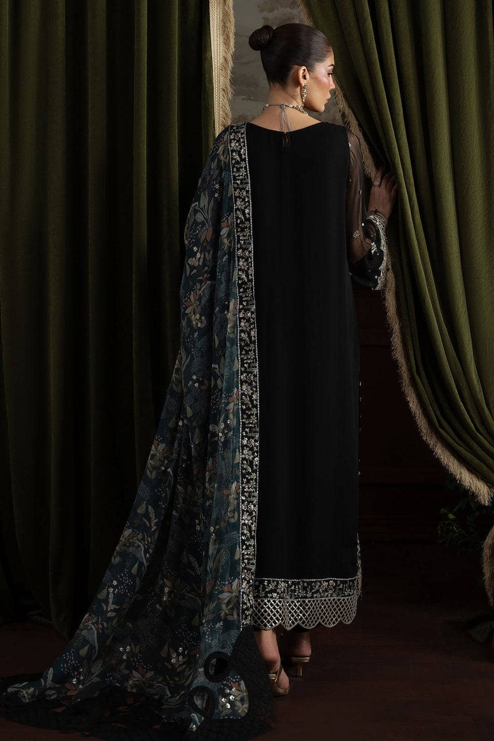 Woman wearing a black dress with a long, patterned scarf draped over her shoulders against a dark curtain background.