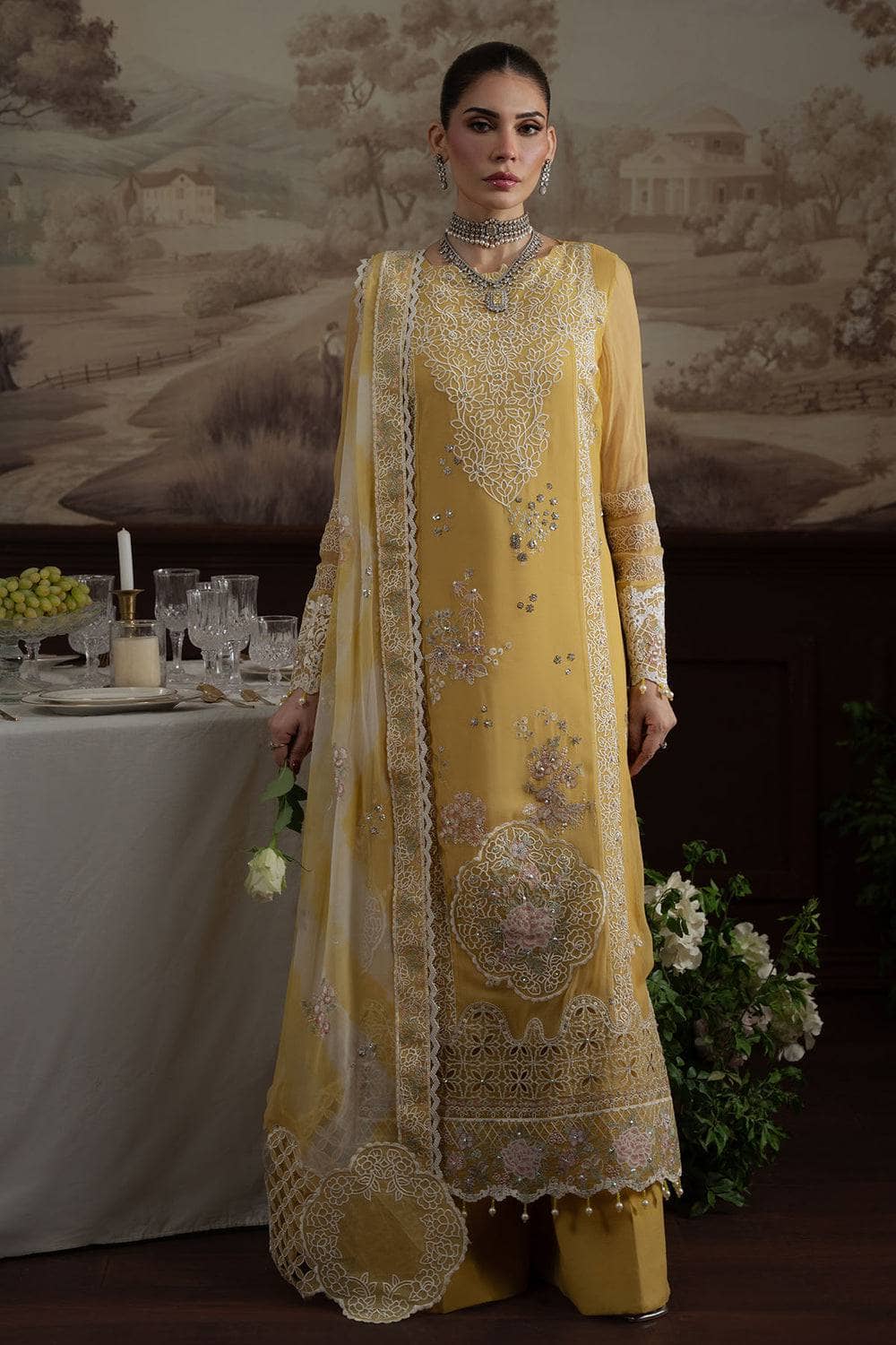 Woman in a yellow embroidered traditional outfit standing in front of a decorated table with a scenic background.