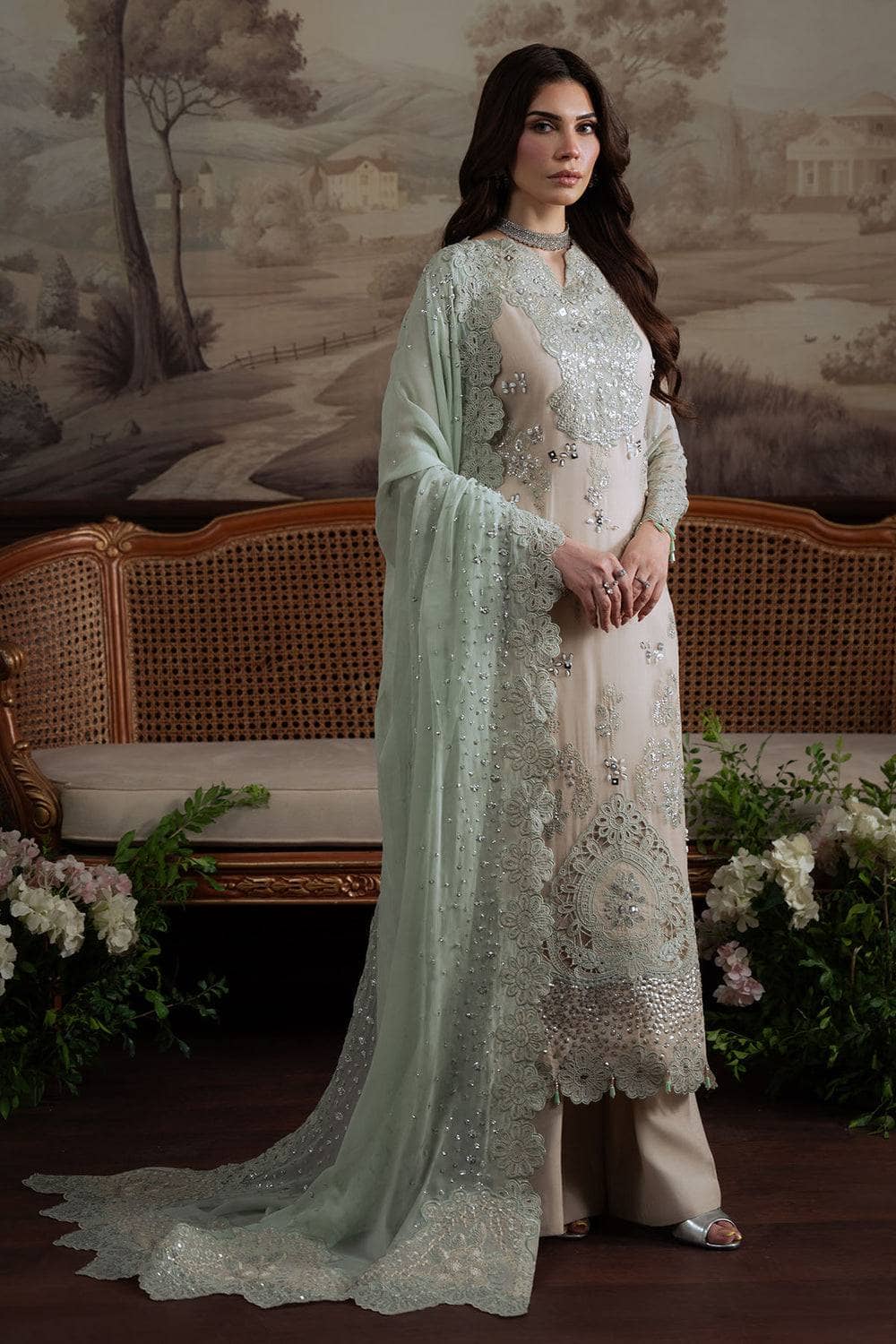 Woman in a light green and white embroidered traditional outfit standing in front of a decorative wall.