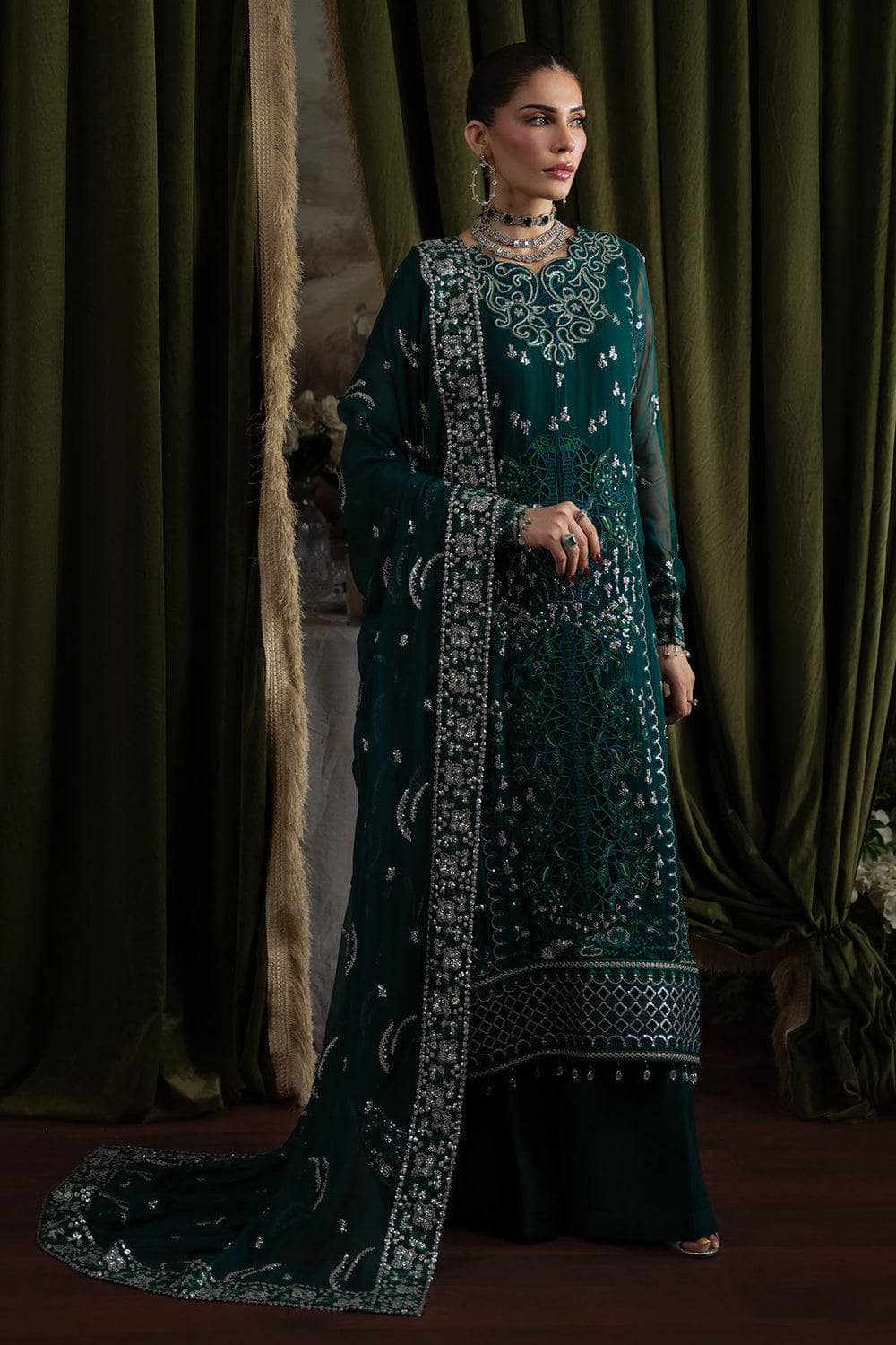 Woman in a dark green embroidered traditional outfit standing against a draped curtain.
