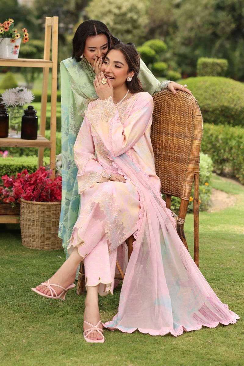 Two women in traditional attire sitting on a grassy area with plants and flowers in the background.