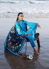 Woman in blue traditional outfit with floral shawl sitting on a beach.