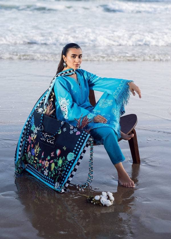 Woman in blue traditional outfit with floral shawl sitting on a beach.