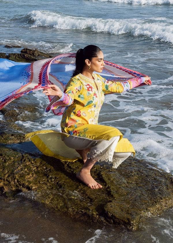 Woman in a yellow outfit with a colorful scarf sitting on a rock by the ocean.