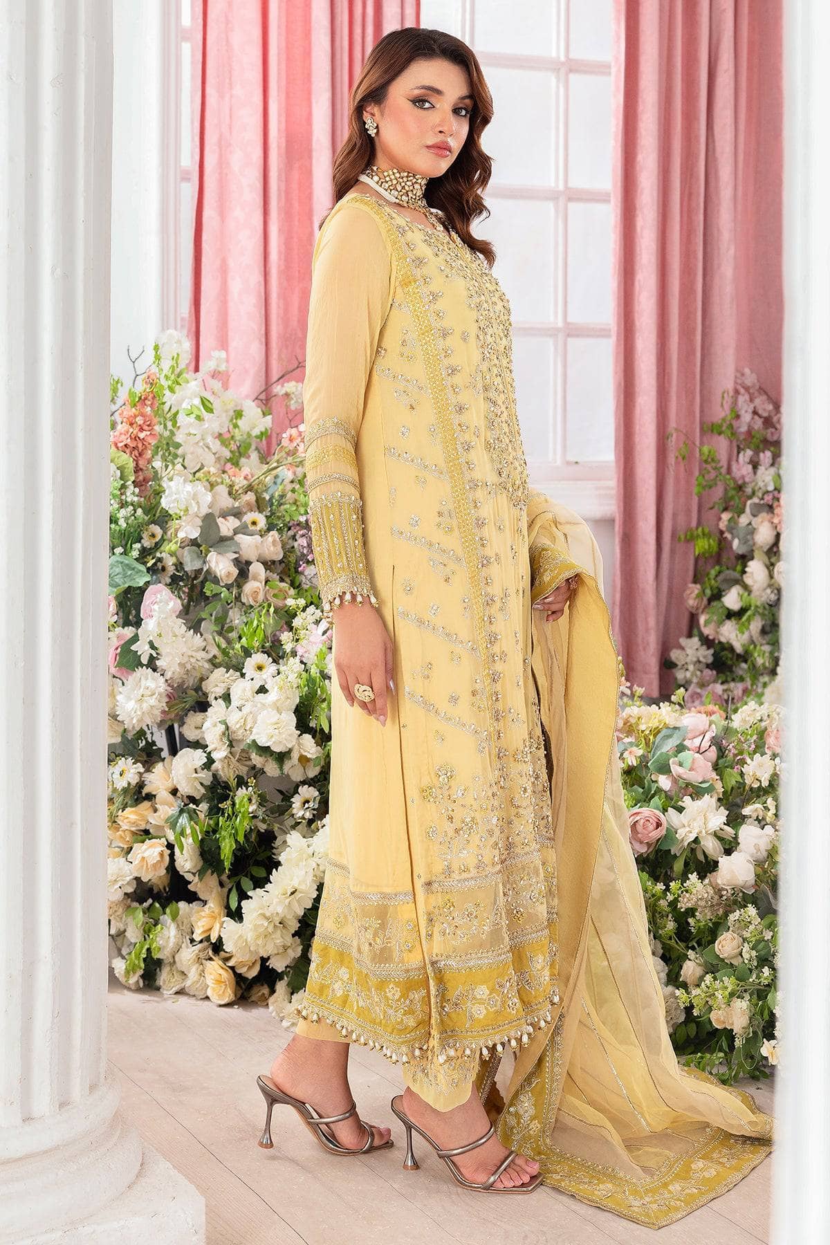 Woman in a yellow embroidered traditional outfit standing in a decorated room with pink curtains and floral arrangements.
