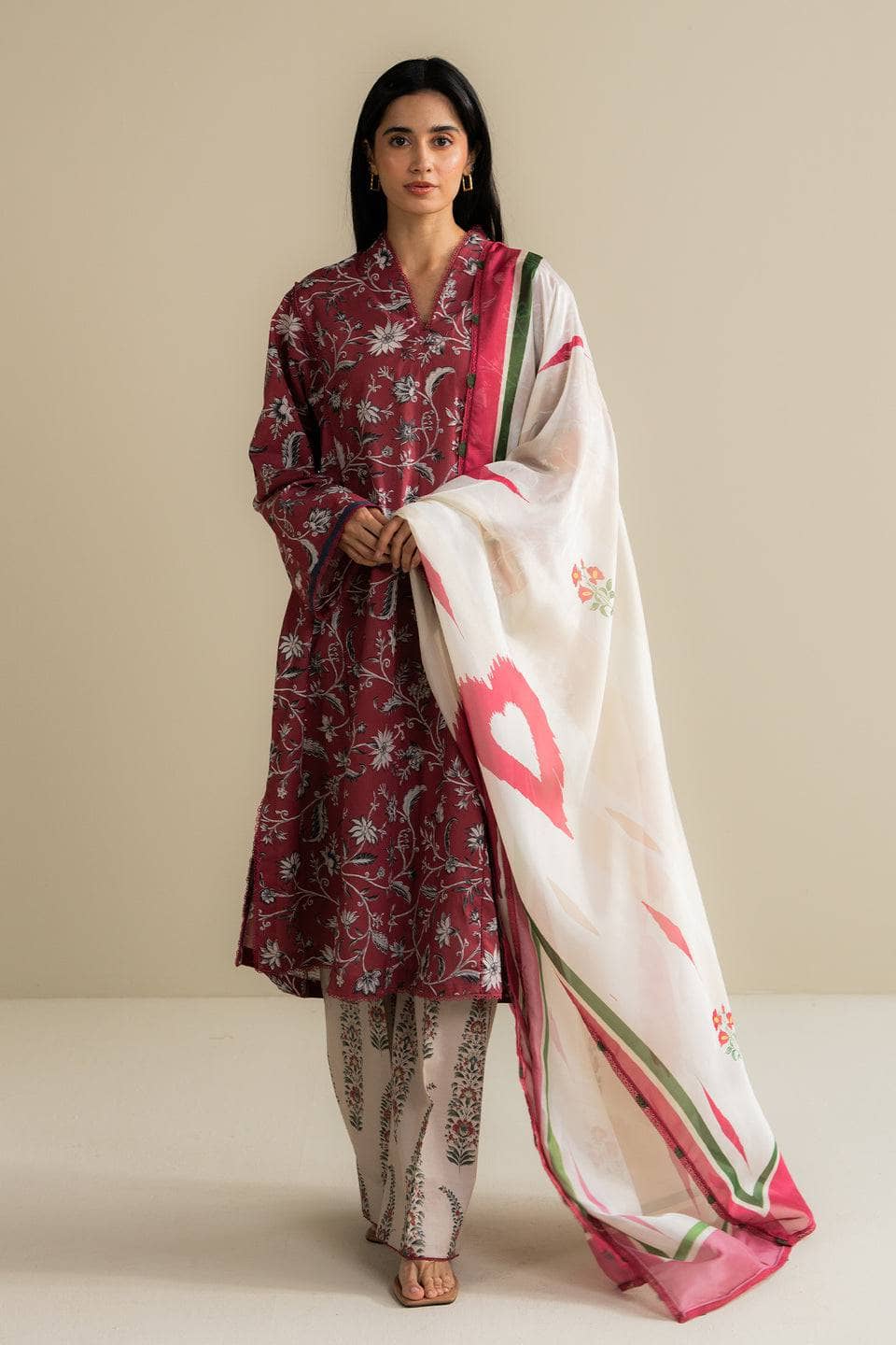Woman wearing a maroon embroidered outfit with a white dupatta featuring pink hearts on a beige background